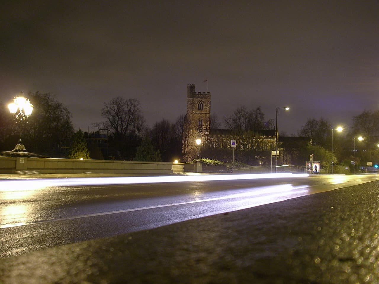 A wet road at night under streetlights, a church tower behind